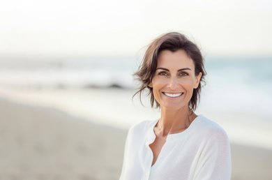 Een lachende vrouw met kort, donkerbruin haar, gekleed in een witte blouse, poseert op een zonnig strand met de zee op de achtergrond.
